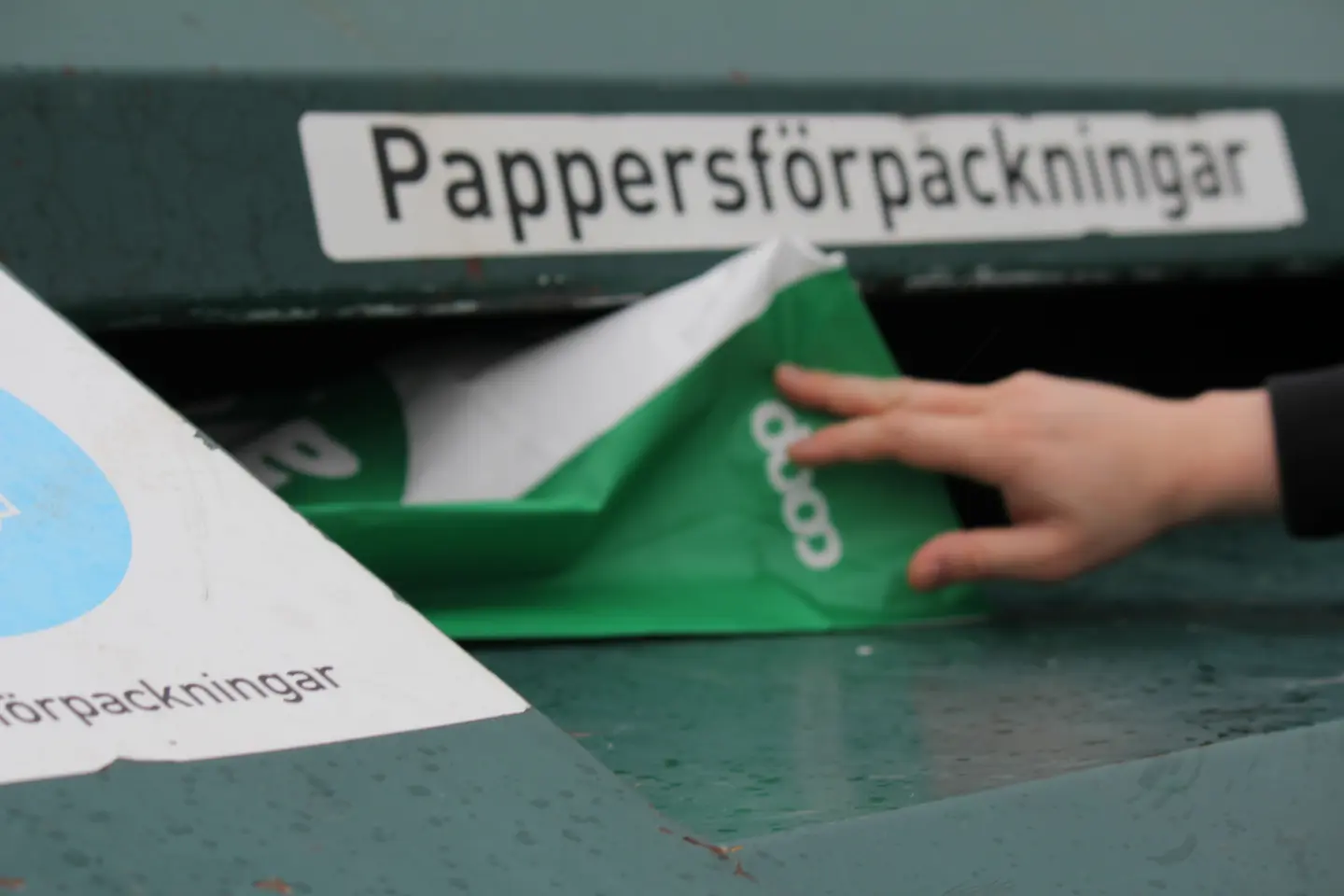 En person slänger en papperspåse i en container märkt "pappersförpackningar" på en återvinningscentral.