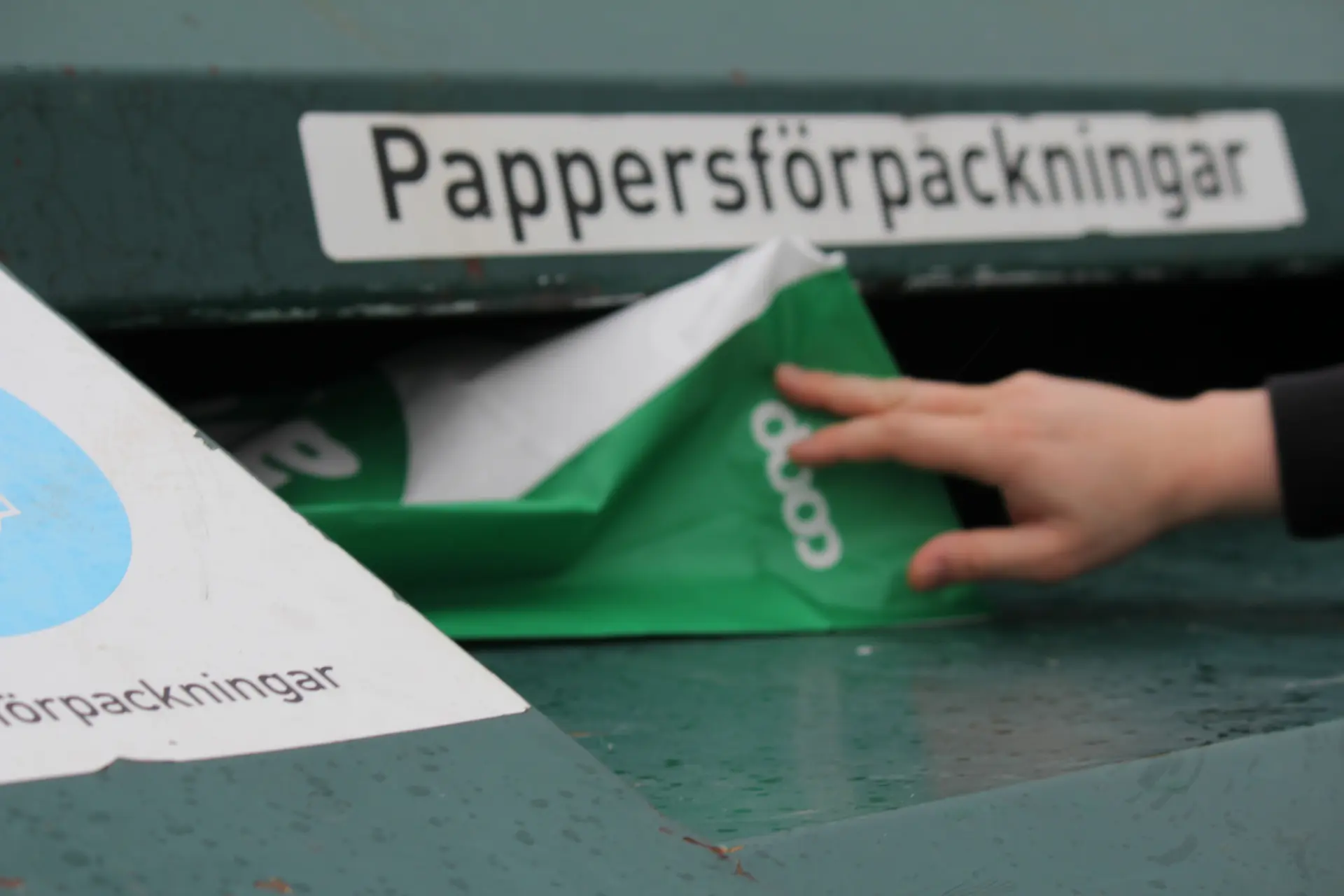 En person slänger en papperspåse i en container märkt "pappersförpackningar" på en återvinningscentral.