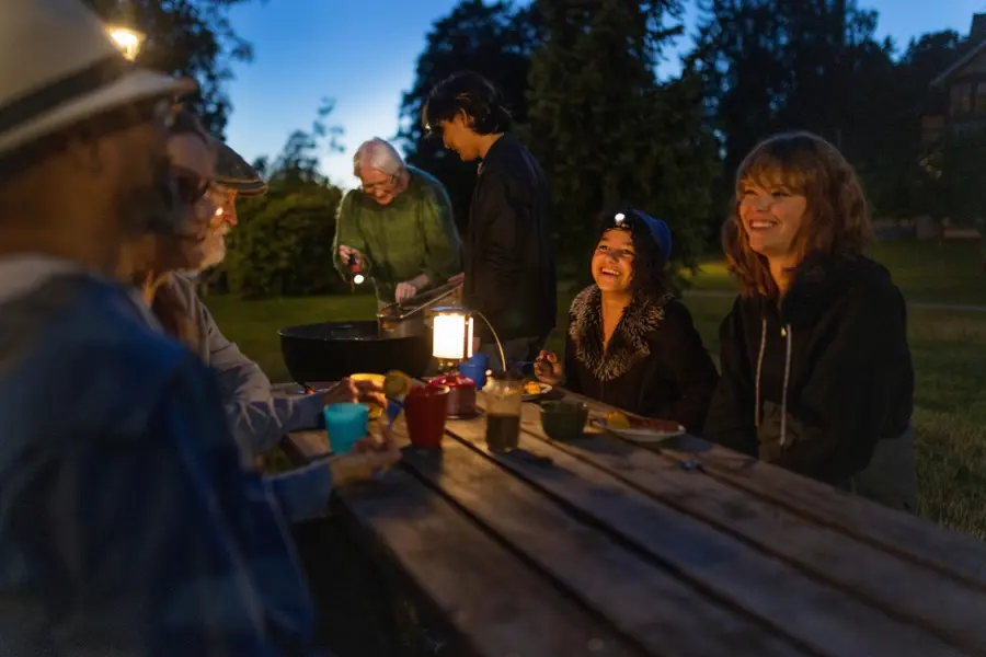 Människor kring matbord utomhus i mörker, i bakgrunden grillas det.