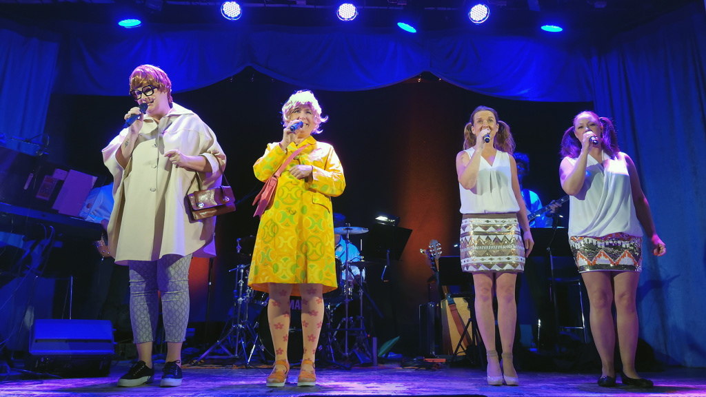 Eva Granlund i gul klänning på scenen tillsammans med tre kvinnor till ur ensemblen.