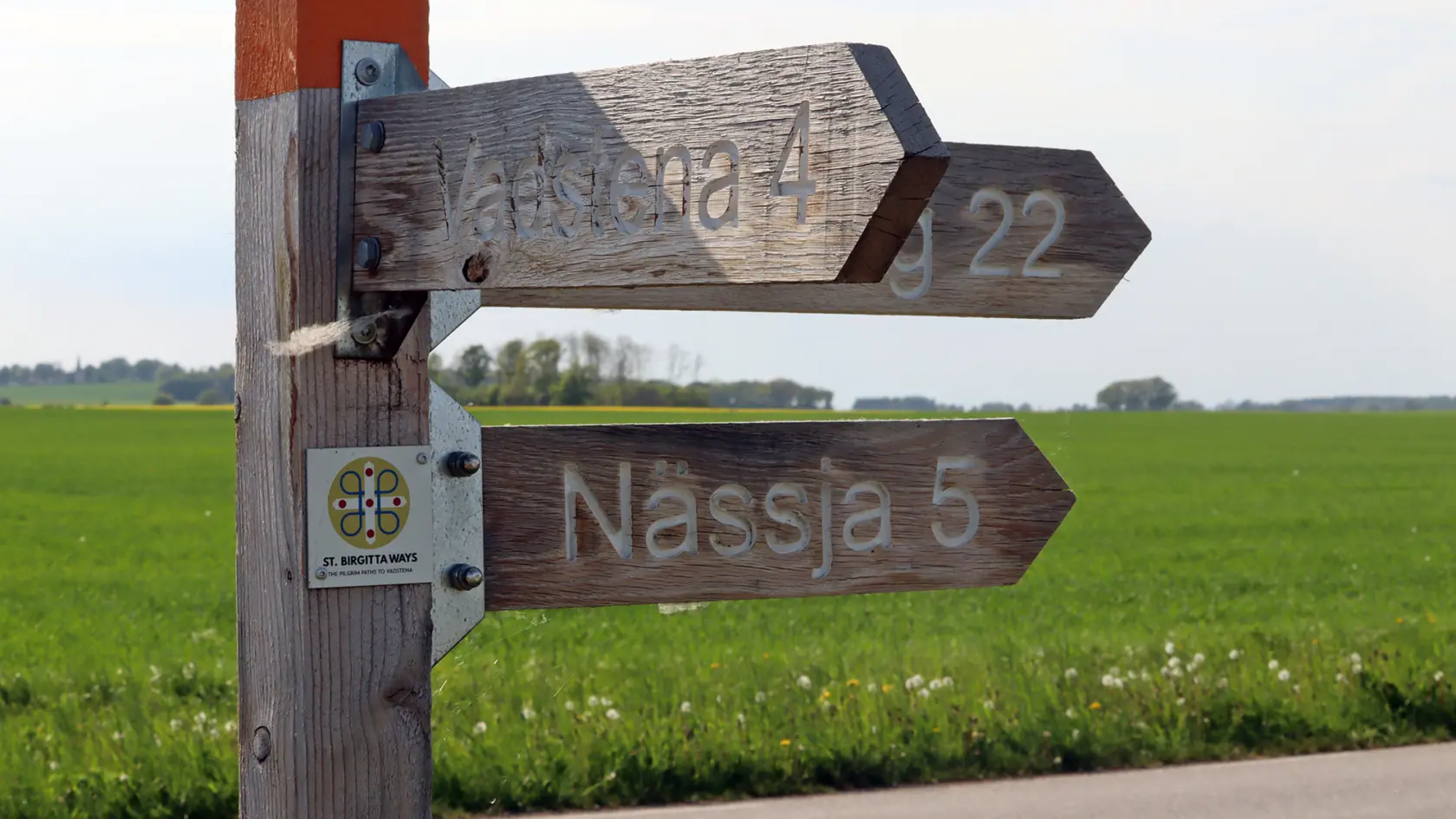 Träskylt på väg vid slätten med pilar mot Vadstena och Nässja