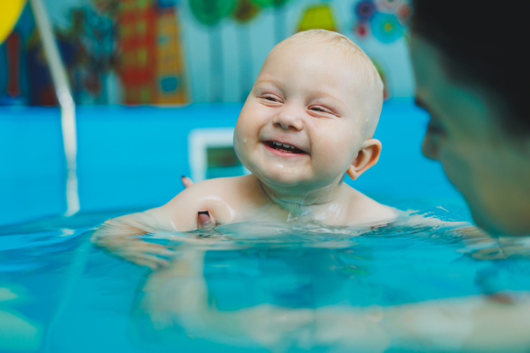 Skrattande baby hålls av mamma i bassäng.