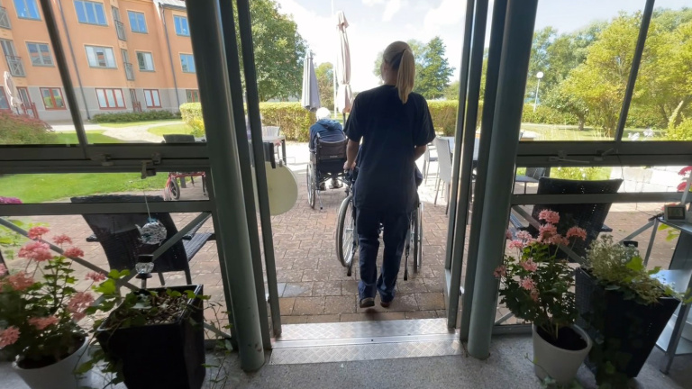 Vårdanställd skjuter en rullstol ut genom en dörr.